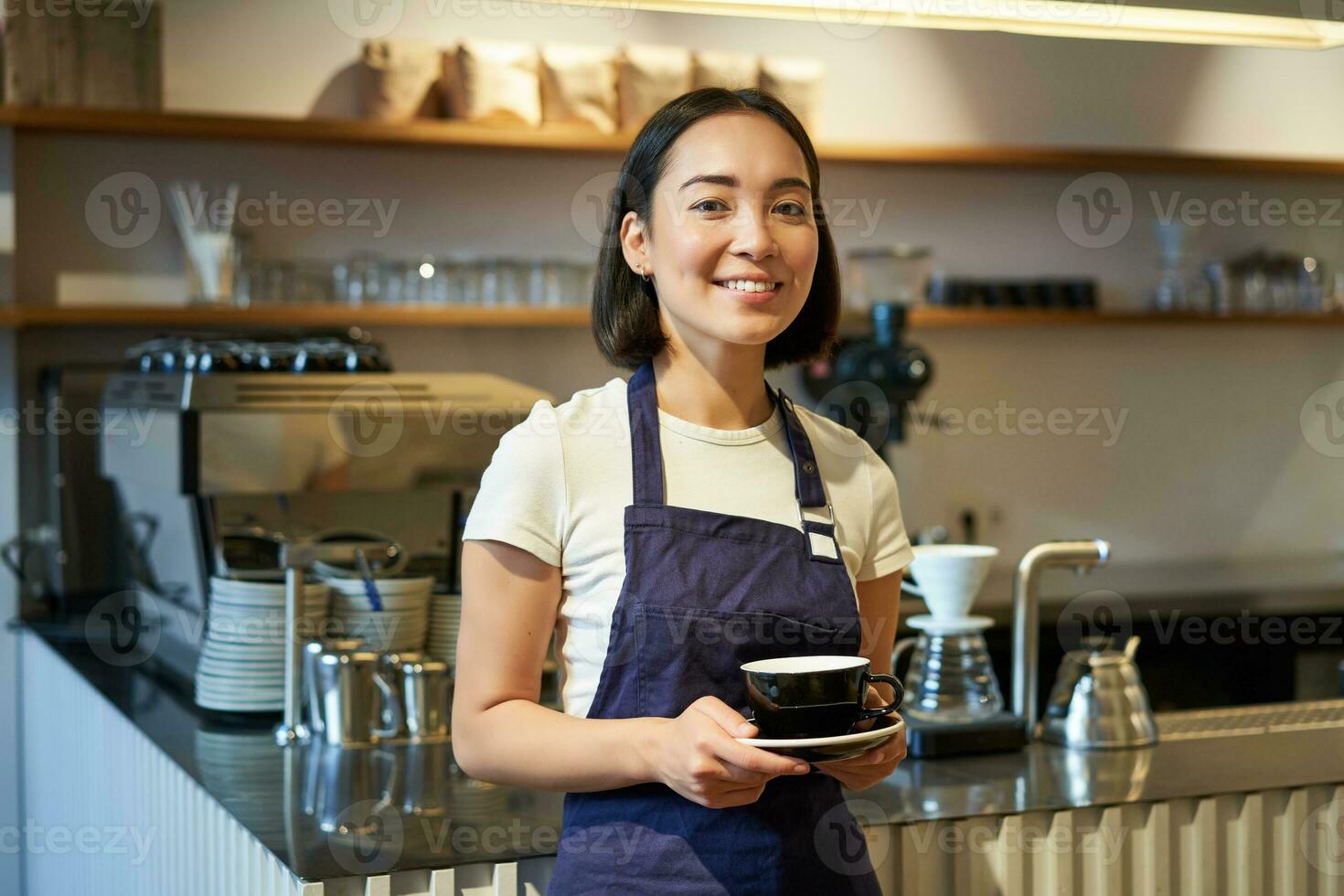 porträtt av asiatisk leende kvinna Barista, bär Kafé förkläde och innehav kopp av kaffe, tar klient beställa till de tabell, tjänande drycker foto