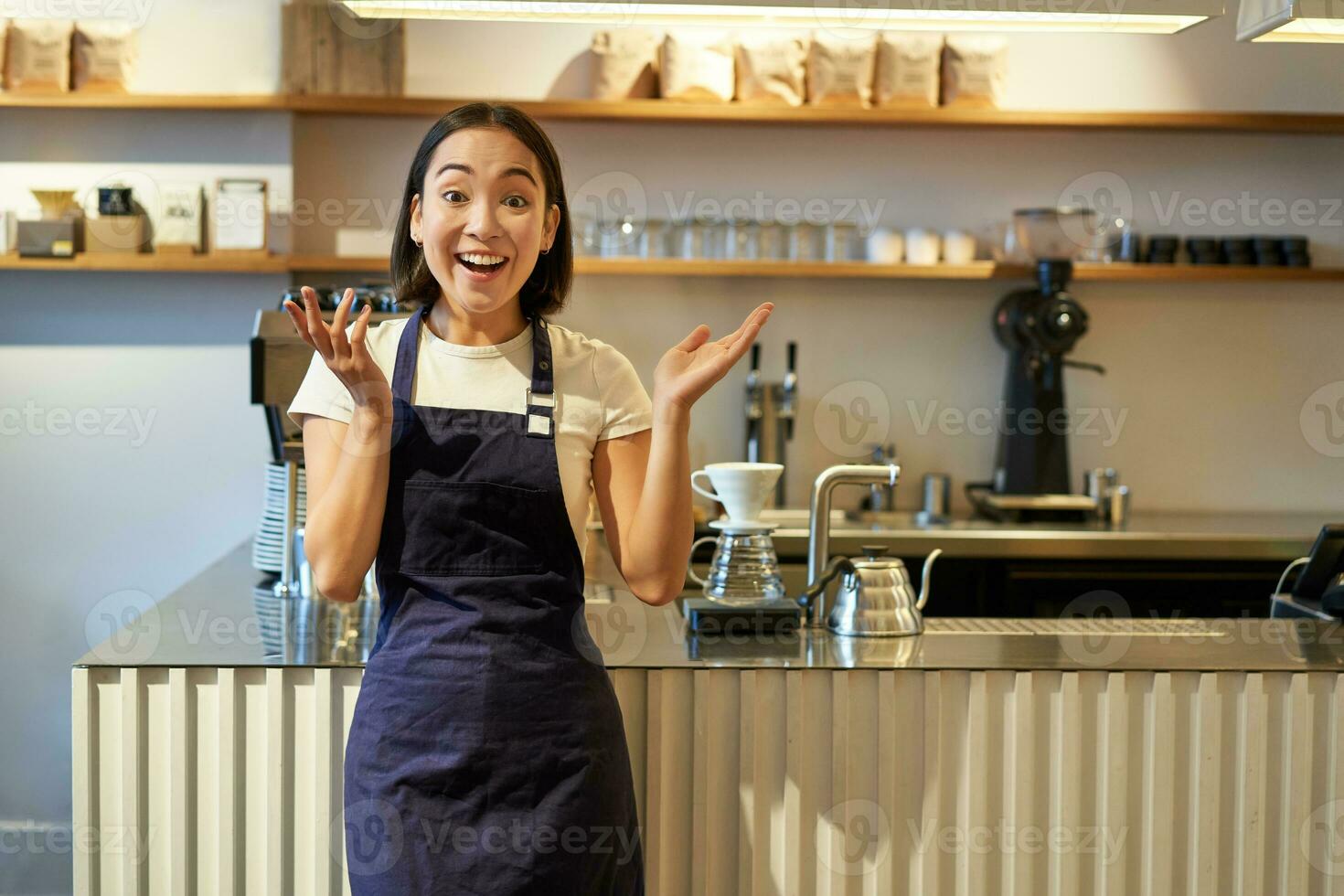entusiastisk ung asiatisk kvinna, Barista i förkläde, ser förvånad, klappa händer och leende, stående i kaffe affär, arbetssätt i Kafé foto
