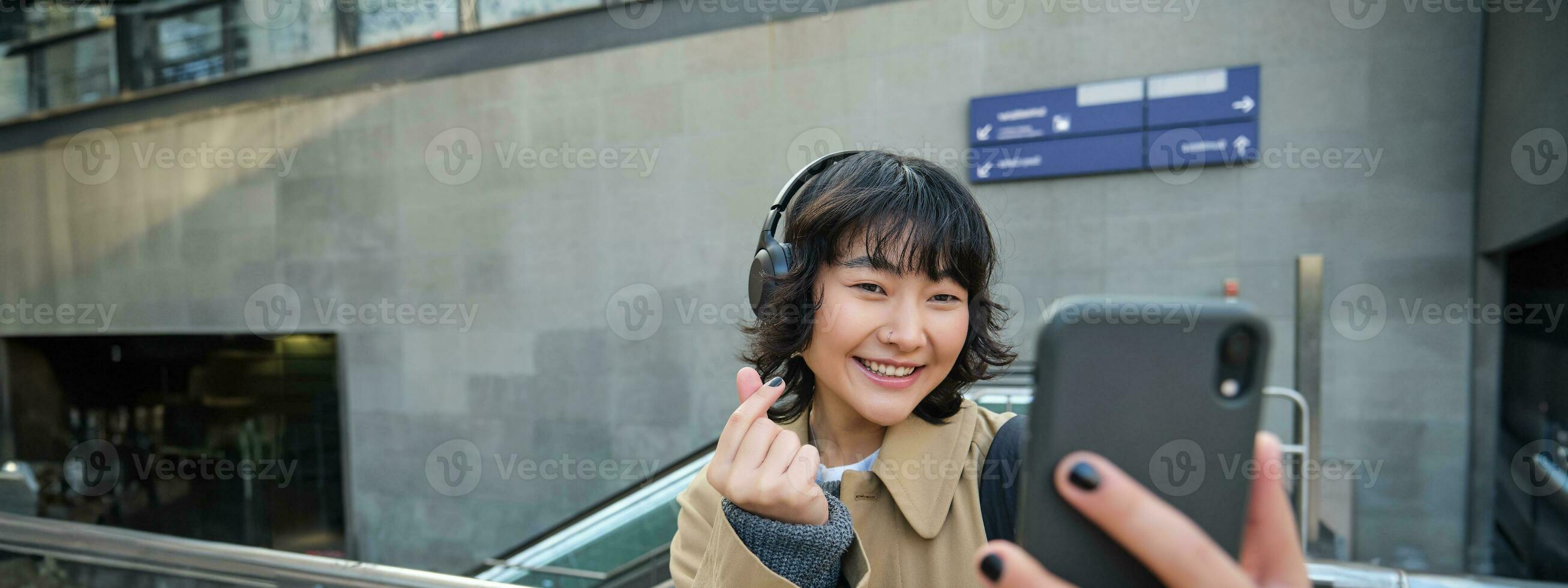 söt och eleganta koreanska flicka, bär hörlurar, tar selfie på smartphone, turist uppgifter video eller gör en Foto, står på gata foto