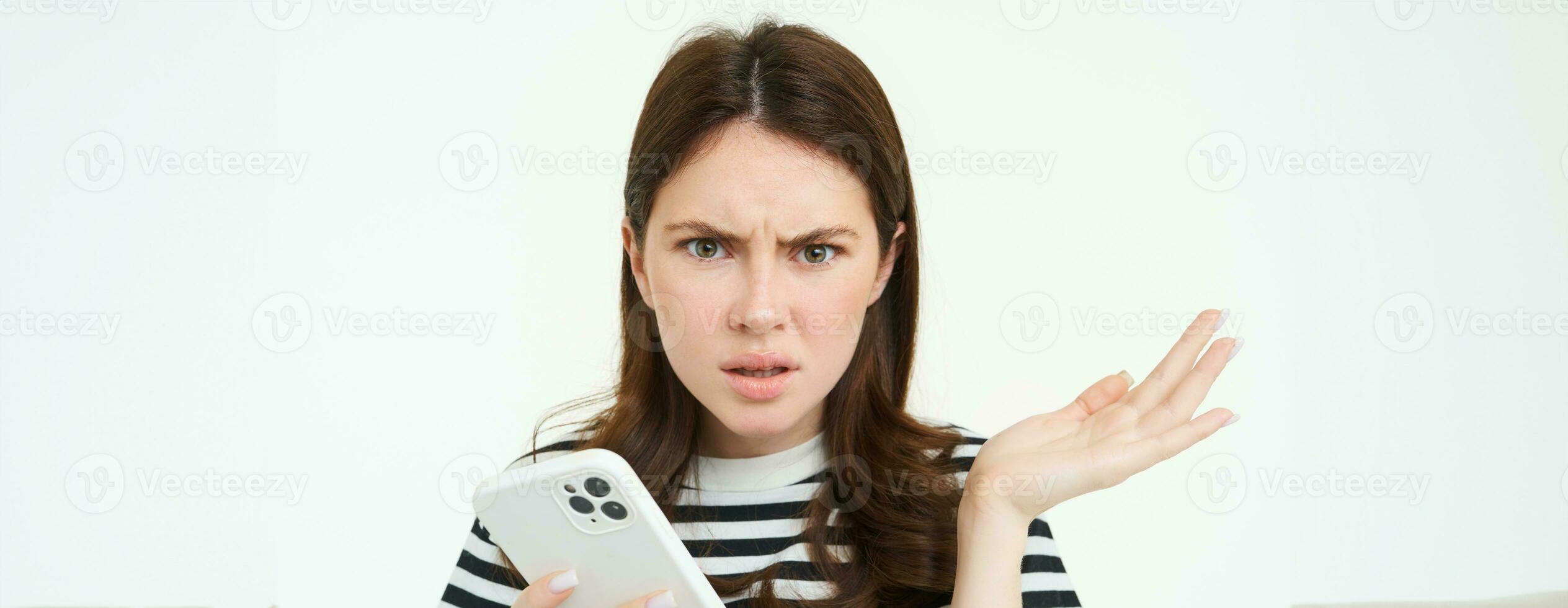 porträtt av förvirrad ung kvinna, utseende frustrerad, axelryckningar axlar, håll mobil telefon i ett hand, vit isolerat bakgrund foto