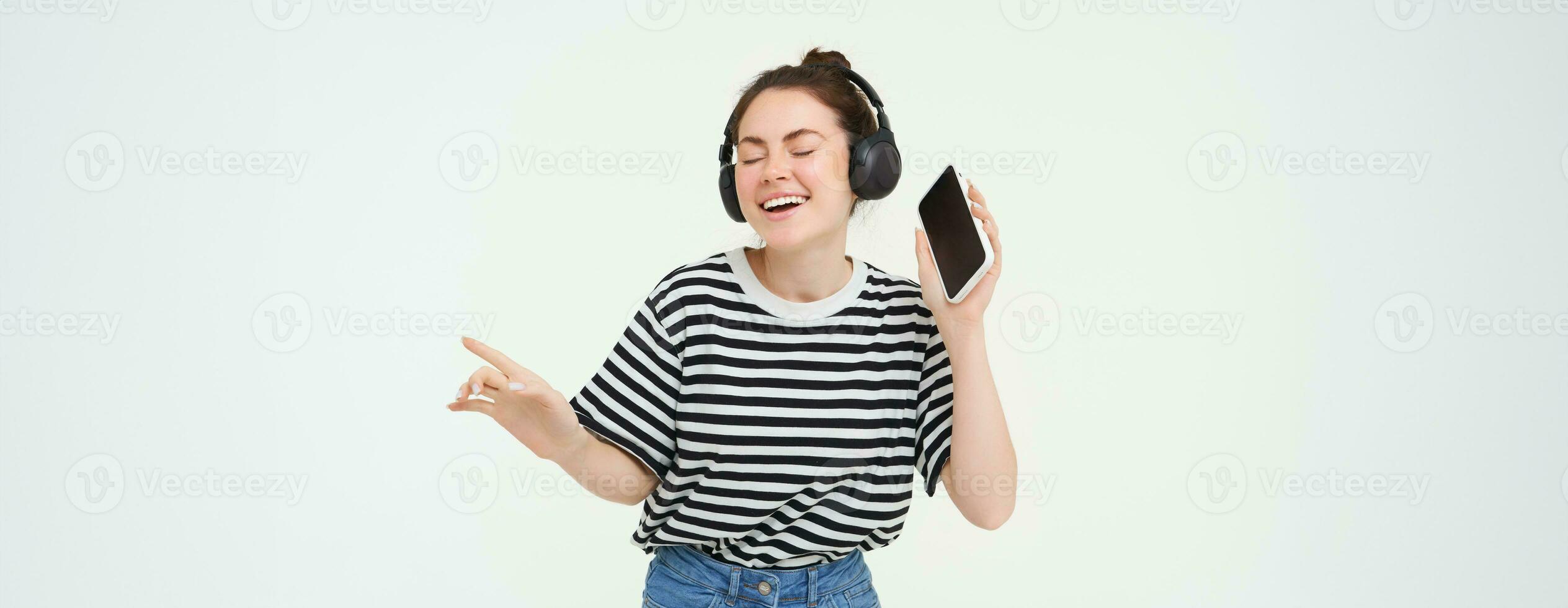 flicka med smartphone dans, lyssnande till musik i trådlös hörlurar, använder sig av mobil telefon strömning app till njut av favorit sånger, isolerat över vit bakgrund foto