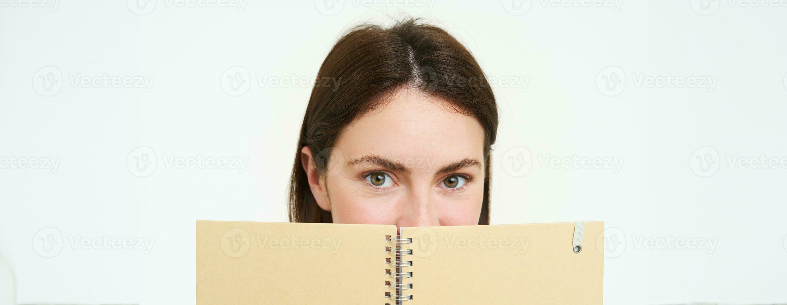 ung kvinna innehar anteckningsbok dagligen planerare Nästa till henne ansikte, skrivning ner läxa, framställning anteckningar, ser omtänksam, stående över vit bakgrund foto