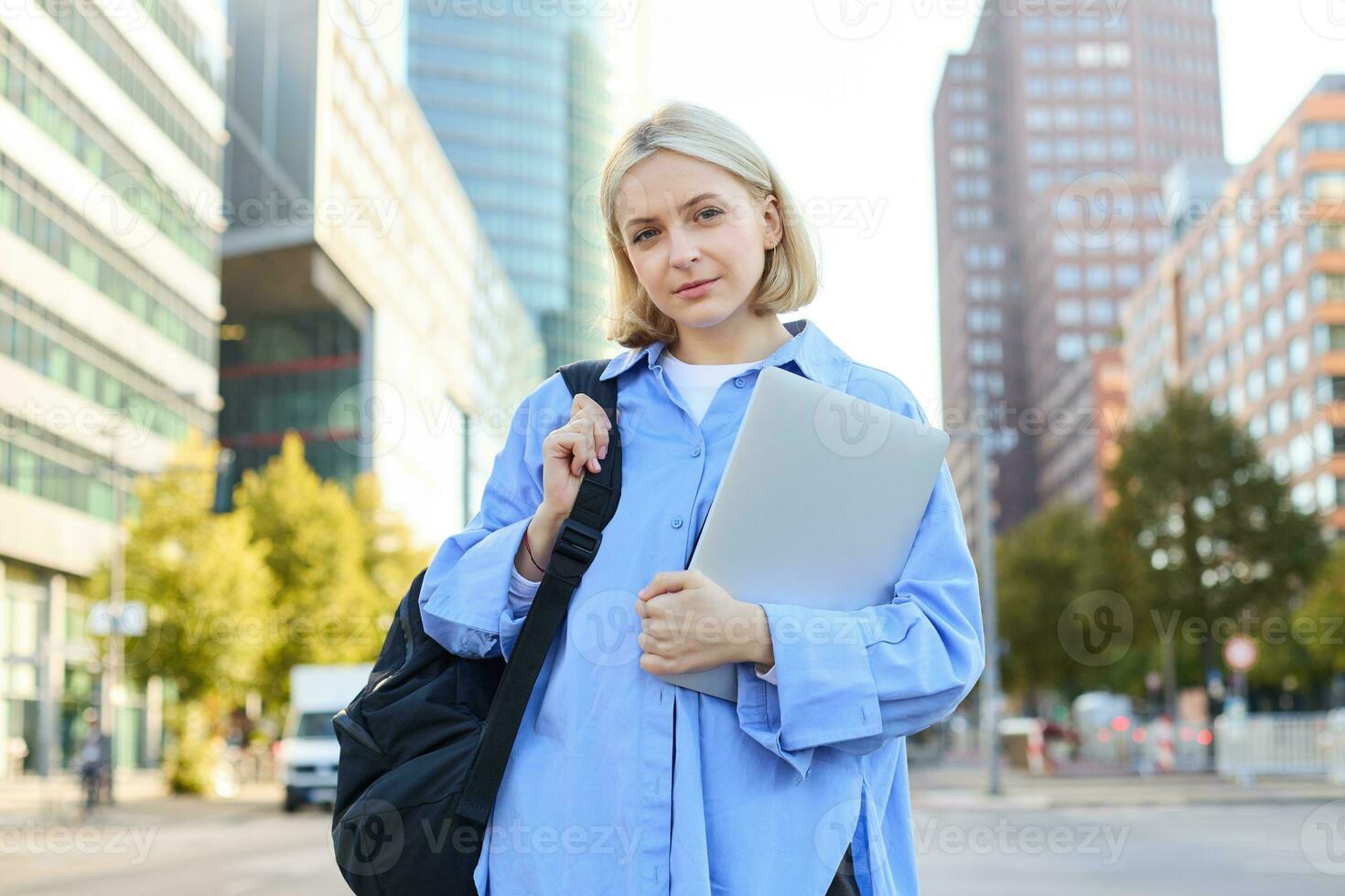 porträtt av ung självsäker kvinna, högskola studerande med ryggsäck och bärbar dator, rubrik till lektion, stående utomhus på tömma gata med stor byggnader Bakom foto