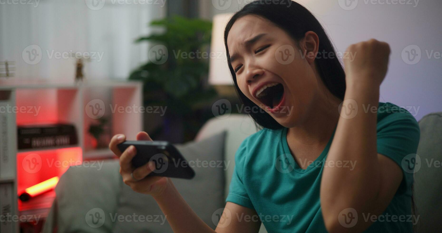 porträtt av ung asiatisk kvinna åtnjuter spelar uppkopplad esport spel på smartphone Sammanträde på soffa i de levande rum på hem, spelare livsstil begrepp. foto