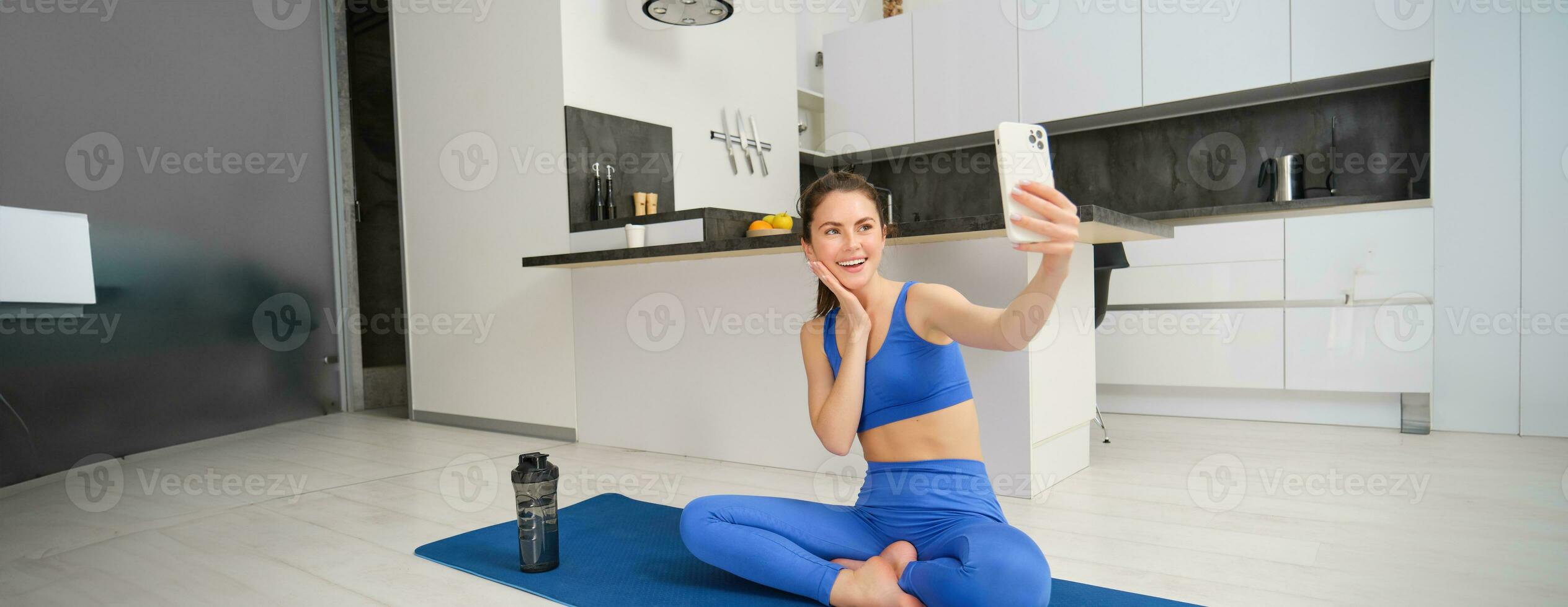 ung leende kvinna, vältränad flicka håller på med träna från Hem, sitter på yoga matta med smartphone, tar selfie, uppgifter sport vlog inomhus foto