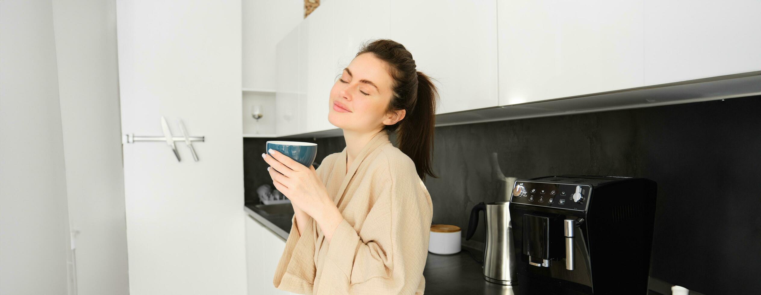 porträtt av kvinna stående nära kaffe maskin, framställning morgon- kopp av cappuccino, stående i kök, bär morgonrock foto