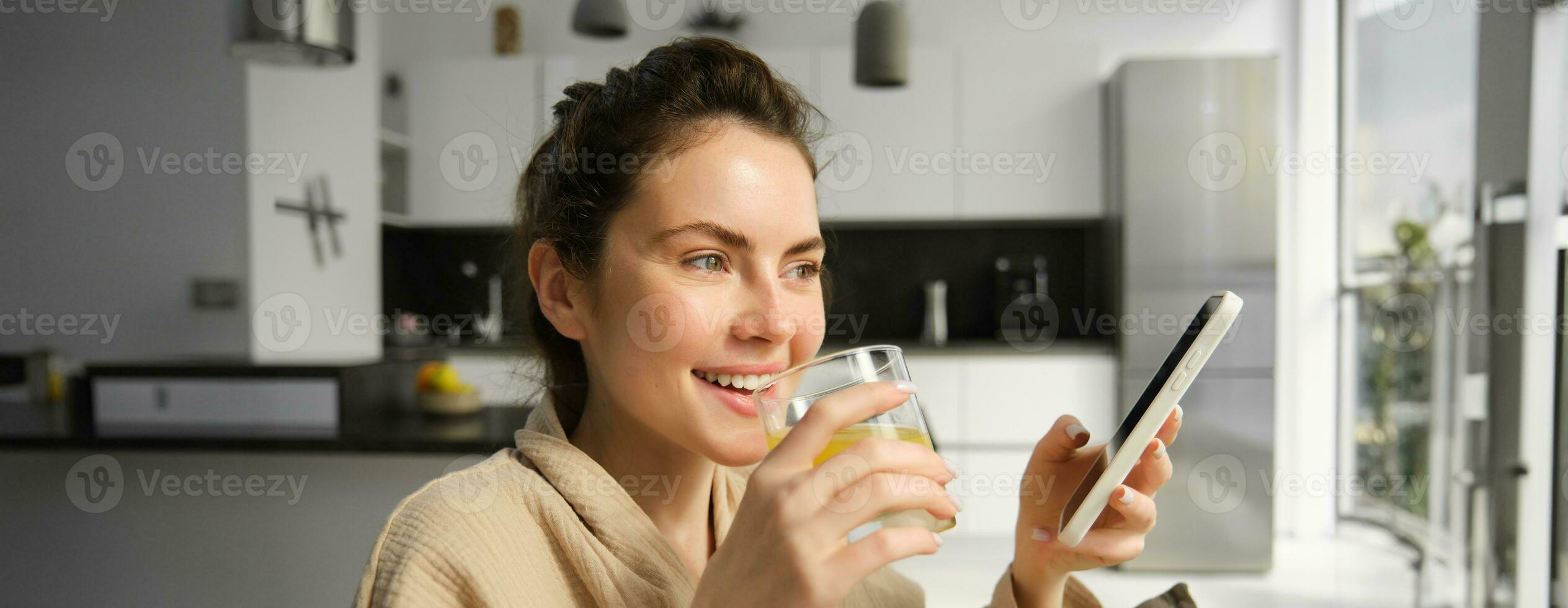 stänga upp porträtt av skrattande, leende ung kvinna åtnjuter morgon, dricka orange juice och innehav smartphone, läsning Nyheter på mobil app foto