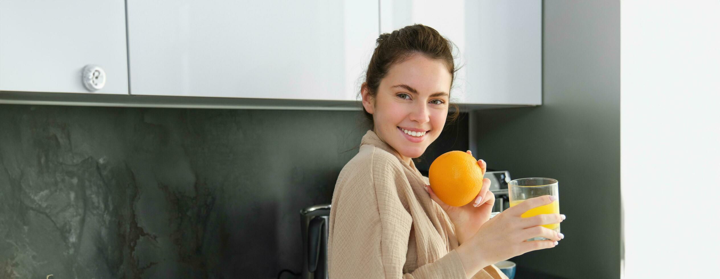 porträtt av Lycklig kvinna i kök, bär morgonrock, dricka orange juice, nyligen pressas dryck, leende och skrattande, mat och dryck begrepp foto