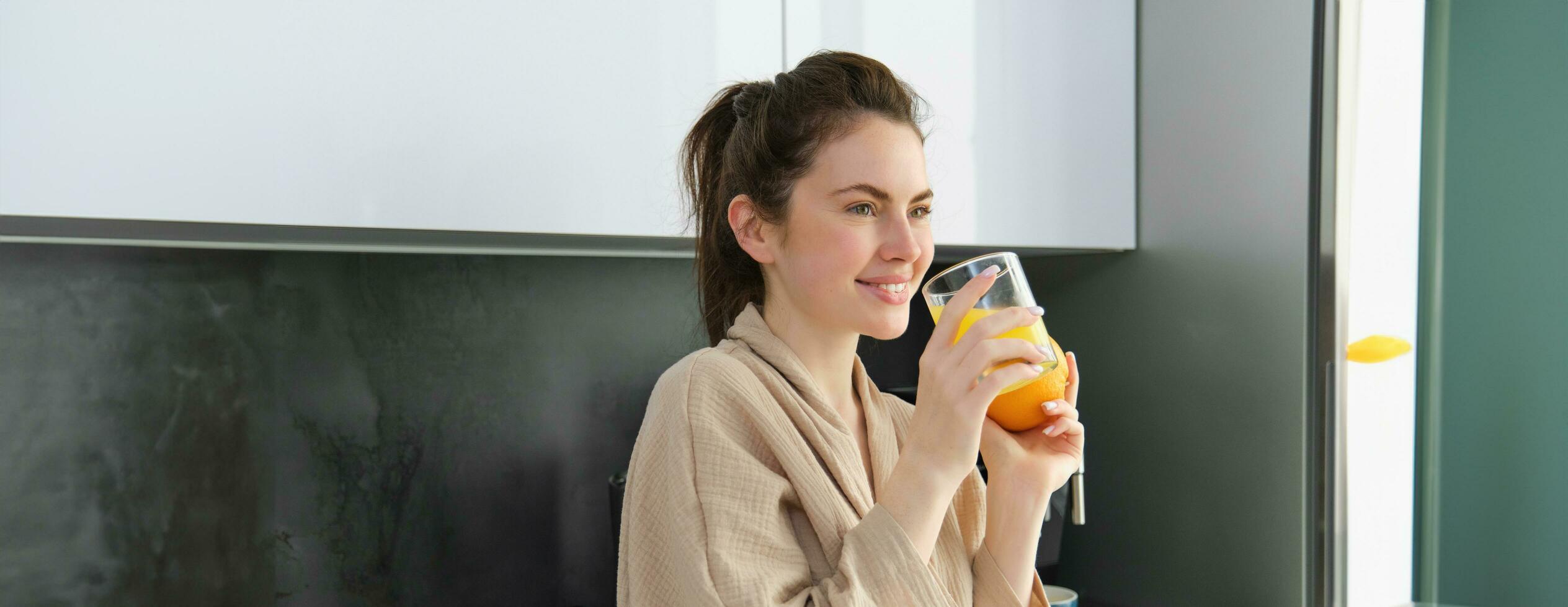 porträtt av snygg kvinna i morgonrock, drycker orange juice från glas och ler, börjar henne dag med friska vana foto