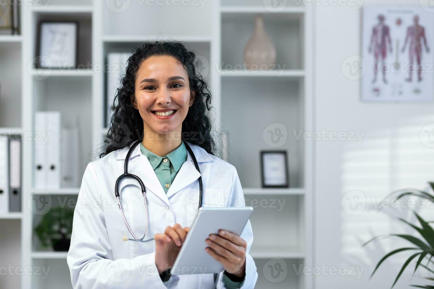 porträtt av framgångsrik leende latinamerikan kvinna läkare inuti medicinsk kontor, kvinna med läsplatta dator stänga upp leende och ser på kamera, arbetssätt i medicinsk klänning medan stående. foto