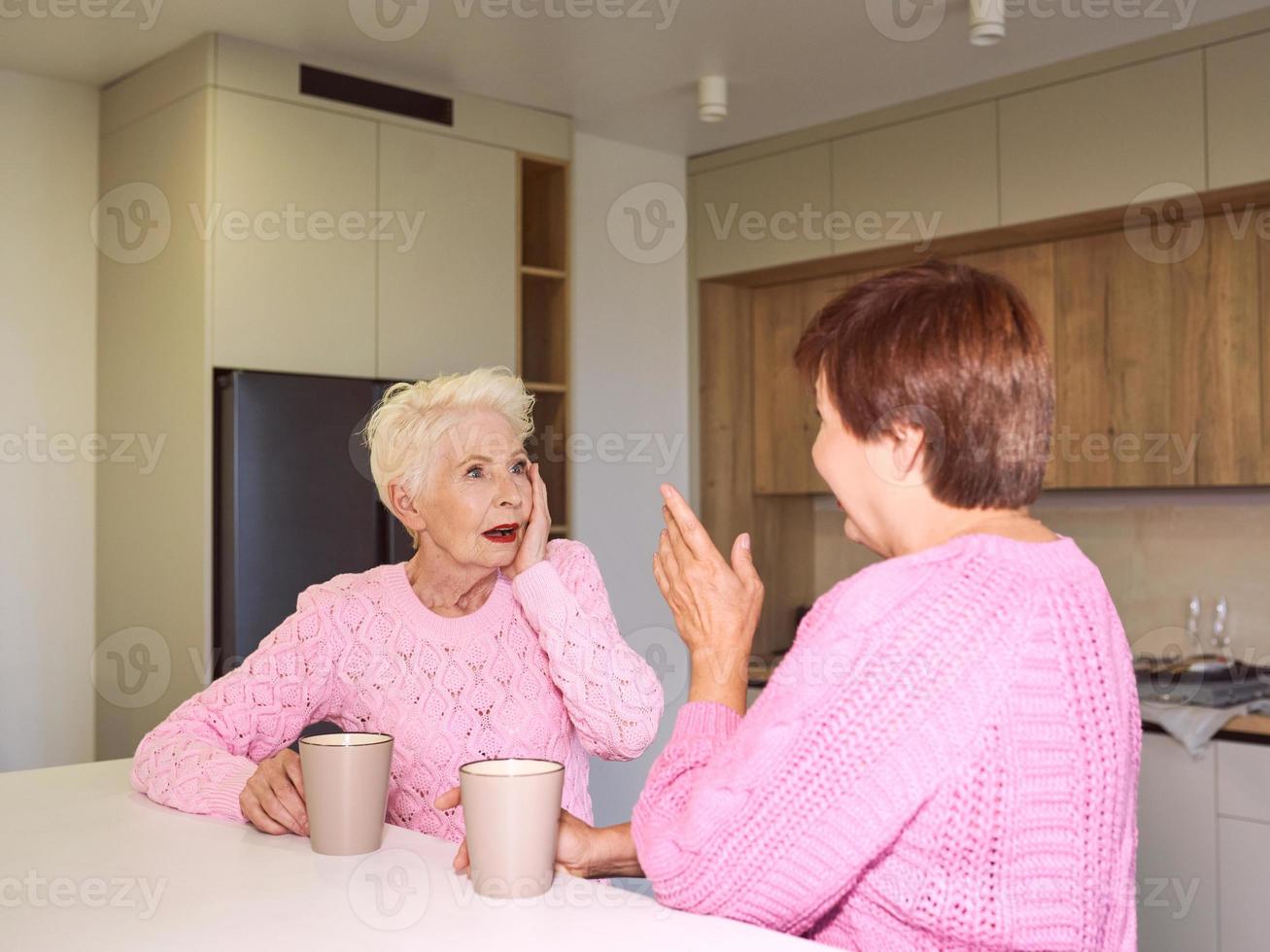 två snygga äldre kvinnor i rosa tröjor som sitter med muggar på moderna köksskvaller. vänskap, prat, skvaller, relationer, nyheter, familjekoncept foto