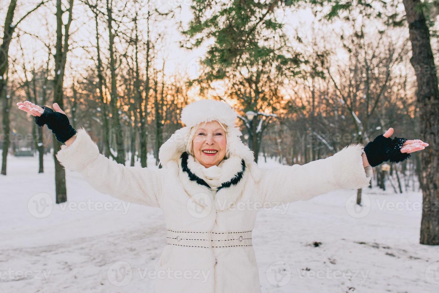 äldre kvinna i vit hatt och kappa njuter av vintern i snöparken. vinter, ålder, säsongskoncept foto