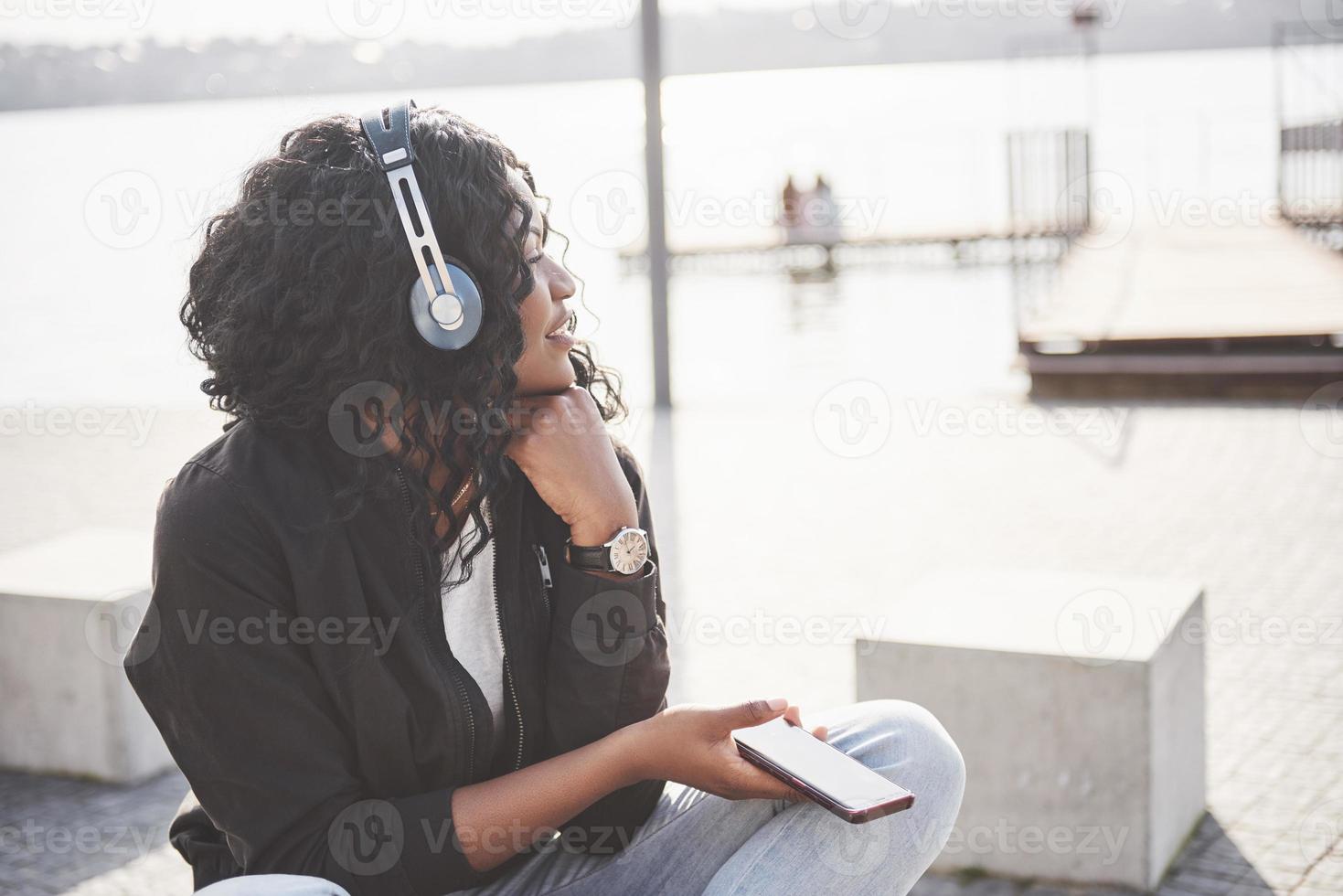 porträtt av en vacker ung söt afroamerikansk flicka som sitter på stranden eller sjön och lyssnar på musik i sina hörlurar foto