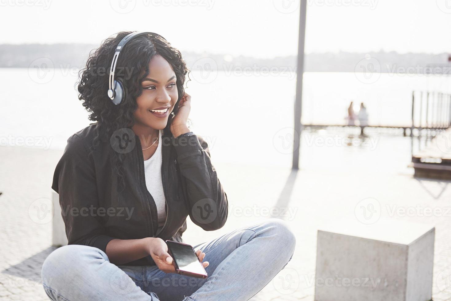 porträtt av en vacker ung söt afroamerikansk flicka som sitter på stranden eller sjön och lyssnar på musik i sina hörlurar foto