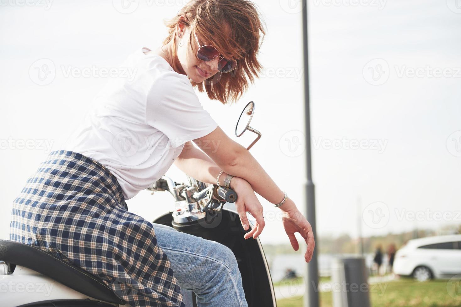 porträtt av en vacker flicka hipster sitter på en svart retro scooter, leende poserar och njut av det varma vårsolen. foto