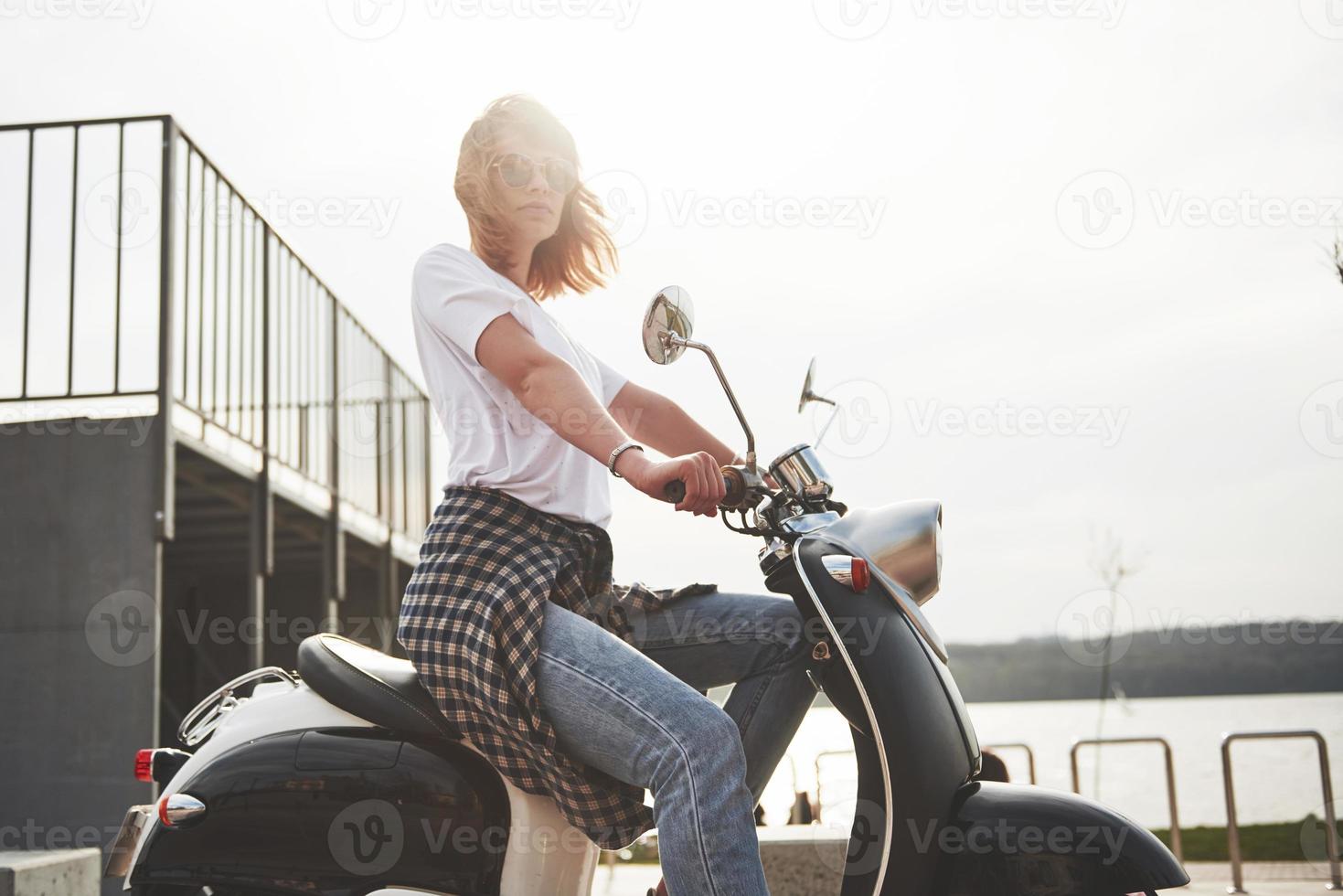 porträtt av en vacker flicka hipster sitter på en svart retro scooter, leende poserar och njut av det varma vårsolen. foto