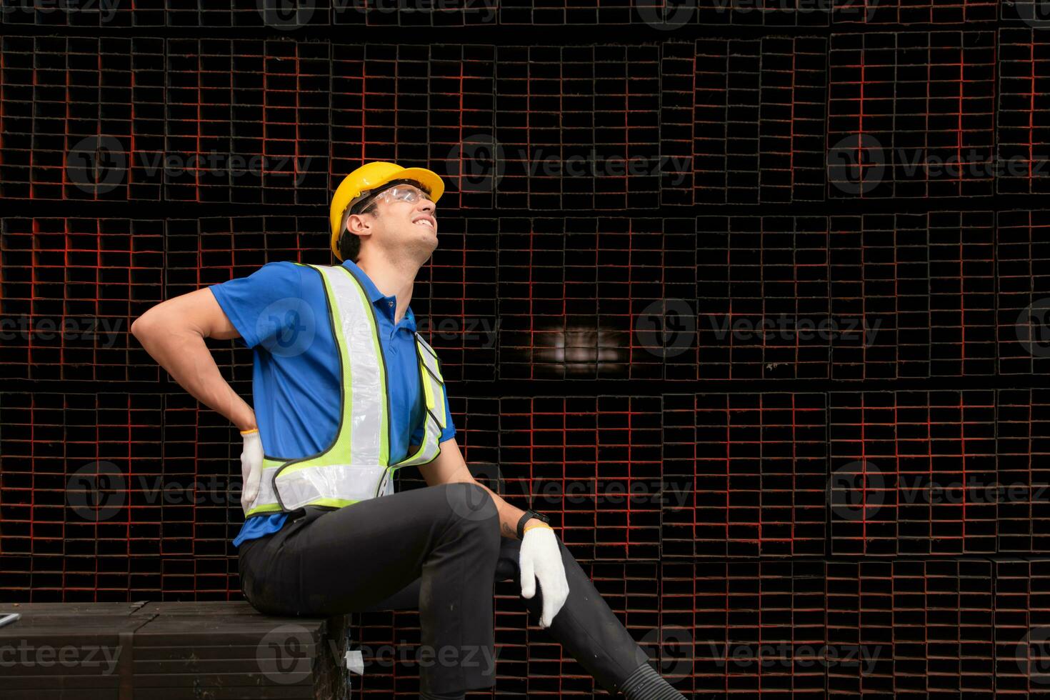 porträtt av en manlig arbetstagare bär en säkerhet väst och hjälm Sammanträde på en stål lastpall på grund av till tillbaka smärta från arbetssätt i en fabrik lyft tung saker. foto