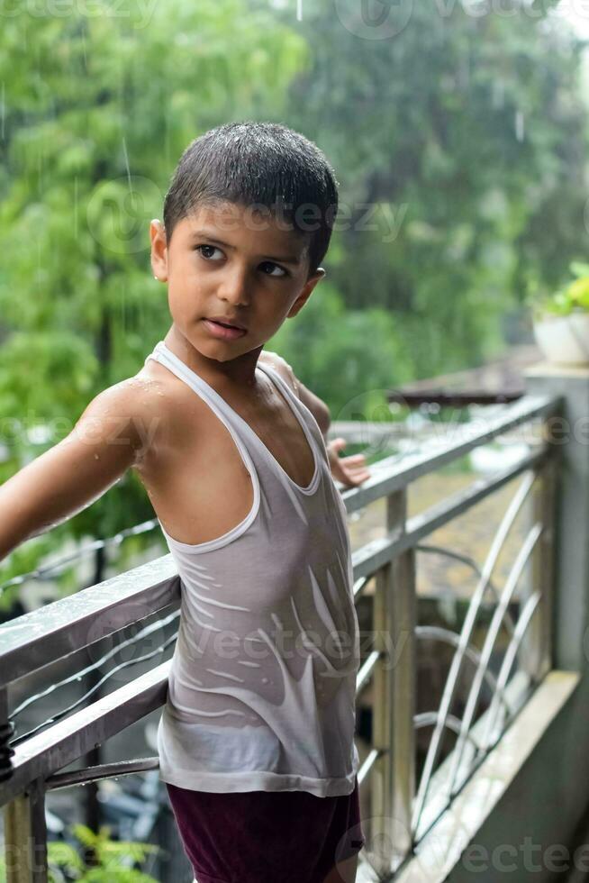 liten unge spelar i sommar regn i hus balkong, indisk smart pojke spelar med regn droppar under monsun regnig säsong, unge spelar i regn foto