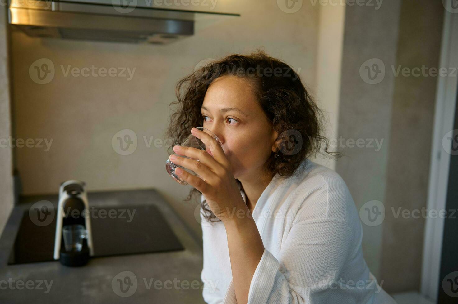 närbild porträtt av en latin amerikan ung vuxen självsäker Söt kvinna i vit morgonrock, dricka vatten i de morgon. foto
