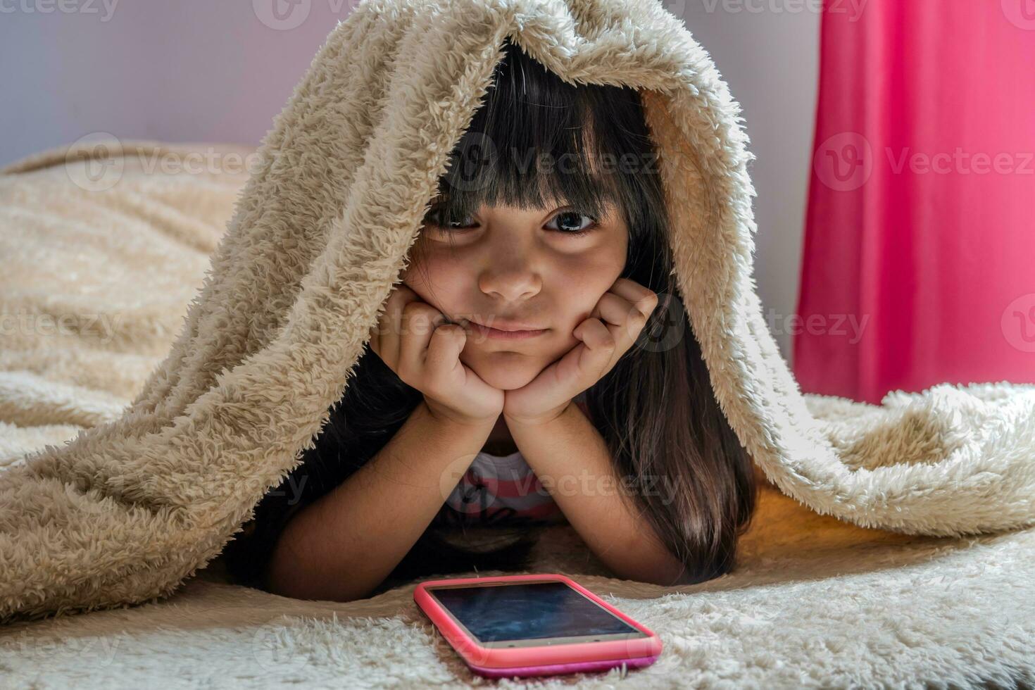 en barn använder sig av en smartphone liggande i säng, spelar spel, tittar på uppkopplad videoklipp, rullning de skärm. barns skärm missbruk. barns rum. foto