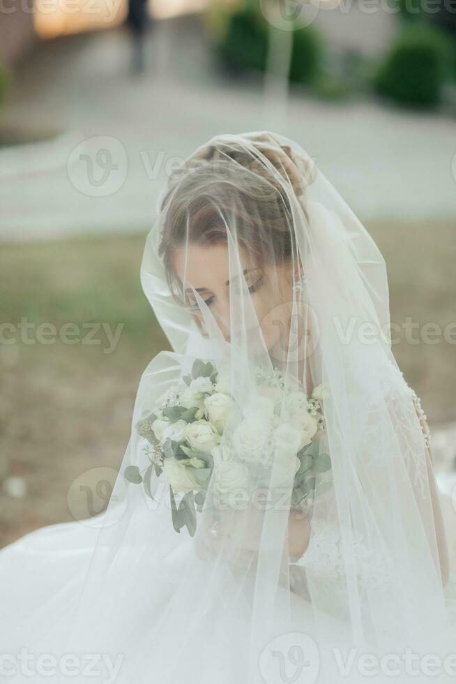 lockigt blond brud i en vit klänning, täckt med en slöja, poser för de kamera med en bukett av rosor. porträtt av de brud. skön smink och hår. bröllop i natur foto