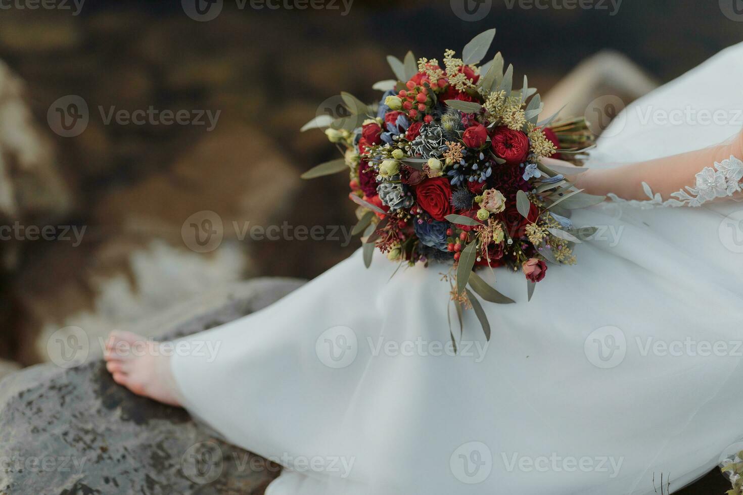 brud på de Bank av de flod, med bar ben Sammanträde på en sten innehav en bukett av olika blommor, röd ro och blå blommor och en lång vit klänning foto