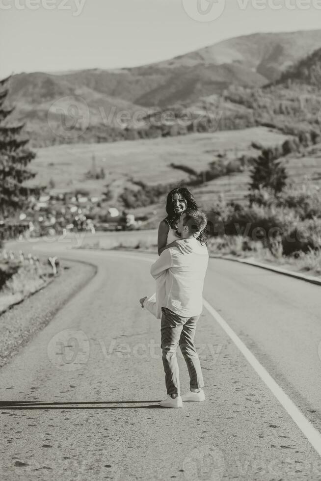 en eleganta brudgum i en vit skjorta och en söt brunett brud i en vit klänning är cirkulerande på ett asfalt väg mot de bakgrund av en skog och berg. svart och vit Foto