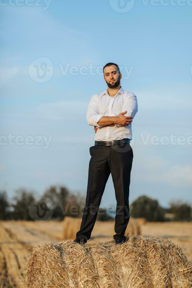 bröllop porträtt Foto. en eleganta brudgum i en vit skjorta poser på en hö bal mot en blå himmel bakgrund. skäggig man. stil. foto