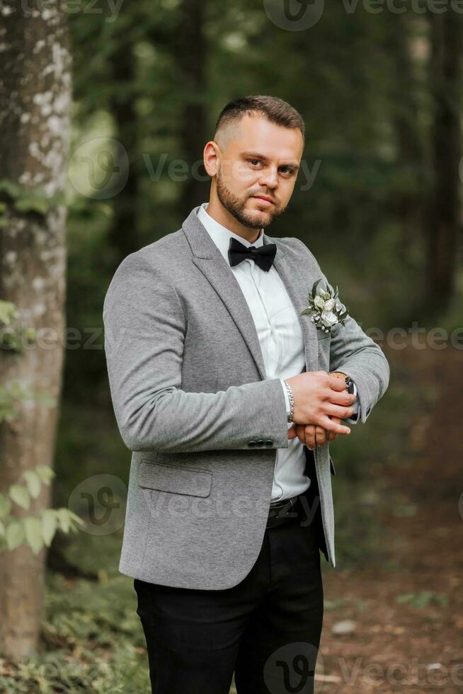 eleganta brudgum justeras hans Kolla på ser in i kamera lins. lyx dyr Kolla på på hand. grå jacka, vit skjorta, vrist stövlar tillverkad av färsk blommor. Foto detaljer