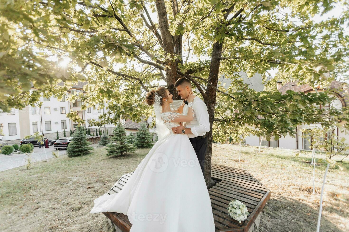 bröllop porträtt. en brudgum i en svart kostym och en blond brud är stående, kramas och kissing under en träd. Foto session i natur. en skön stråle av de Sol i de Foto. skön hår och smink