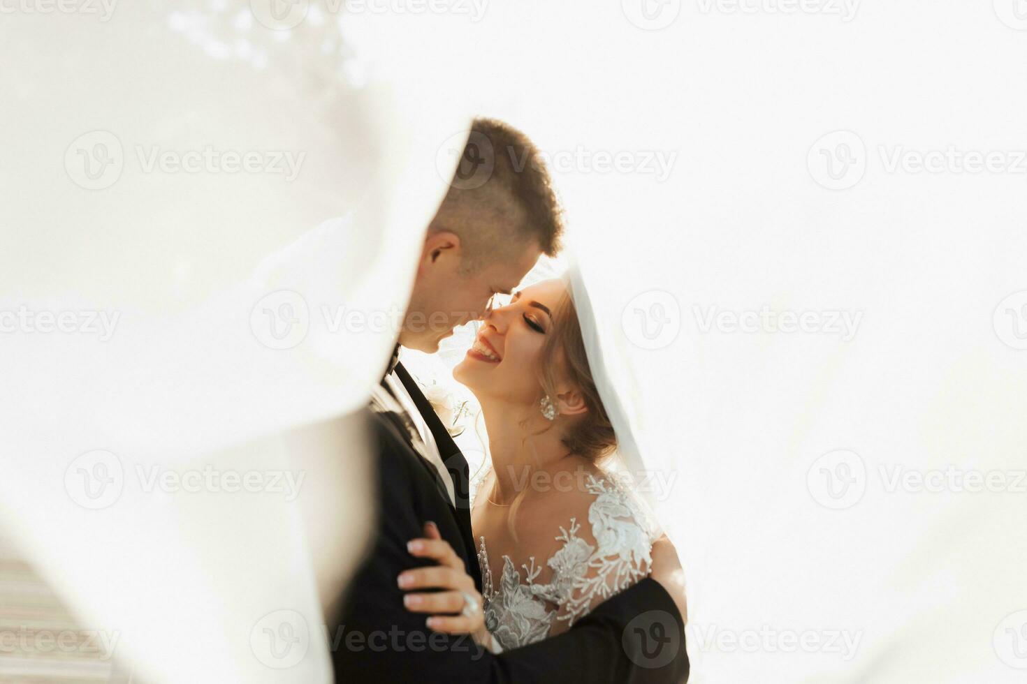 bröllop porträtt. de brudgum i en svart kostym och de blond brud är kramar, insvept i de brudens slöja. lång klänning i de luft. Foto session i natur. skön hår och smink