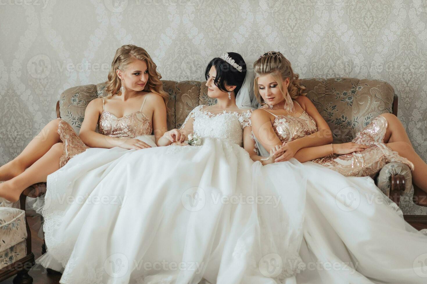 en brunett brud i en vit elegant klänning och krona och henne blond vänner i gyllene klänningar är Sammanträde på de soffa i de brudens rum, leende och Framställ. bröllop porträtt, bröllop Foto. foto