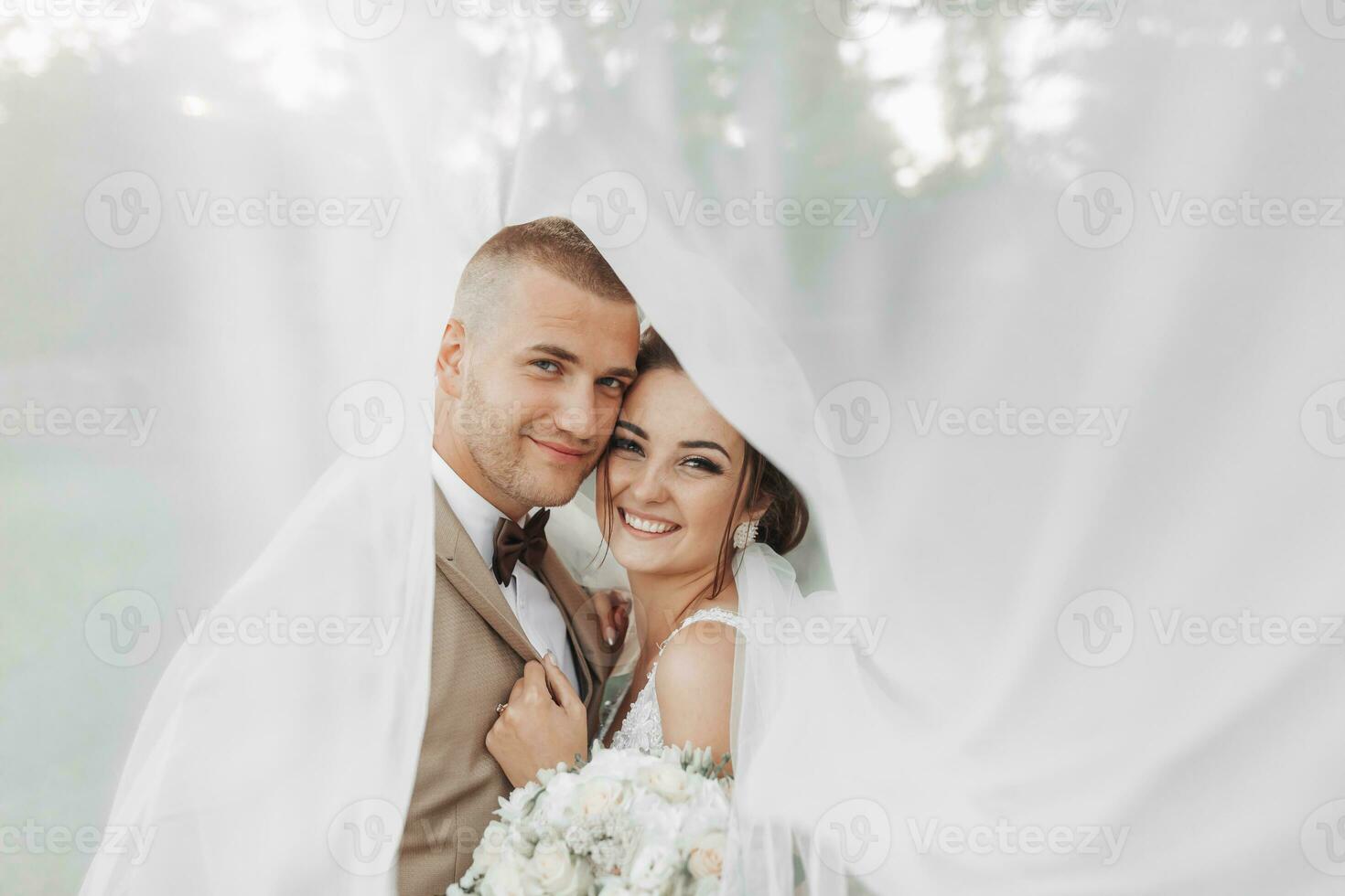 bröllop porträtt av de brud och brudgum. Lycklig nygifta försiktigt kram varje Övrig under de slöja, utgör, se på de kamera och leende. eleganta brudgum. skön ung brud foto