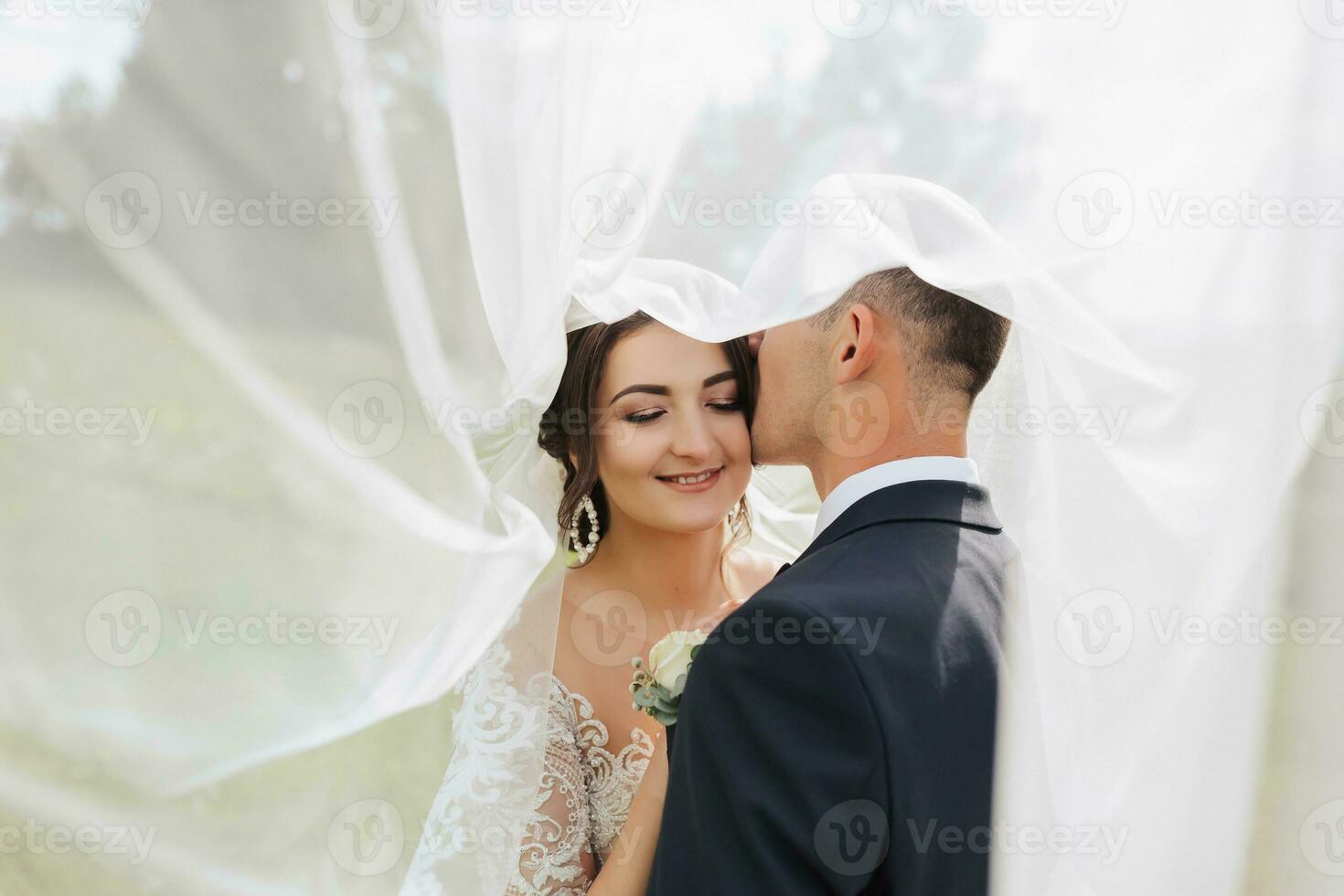 en lockigt hår brunett brud i en vit klänning och en förtäckt brudgum omfamning och kyss. porträtt av de brud och brudgum. skön smink och hår. bröllop i natur foto