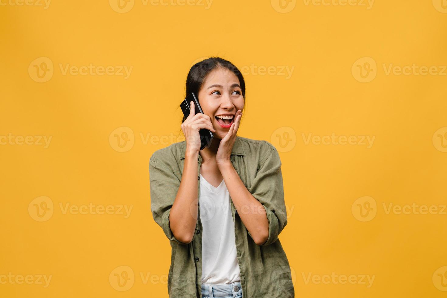ung asiatisk dam pratar i telefon med positivt uttryck, ler brett, klädd i vardagskläder som känner lycka och står isolerad på gul bakgrund. glad förtjusande glad kvinna jublar över framgång. foto