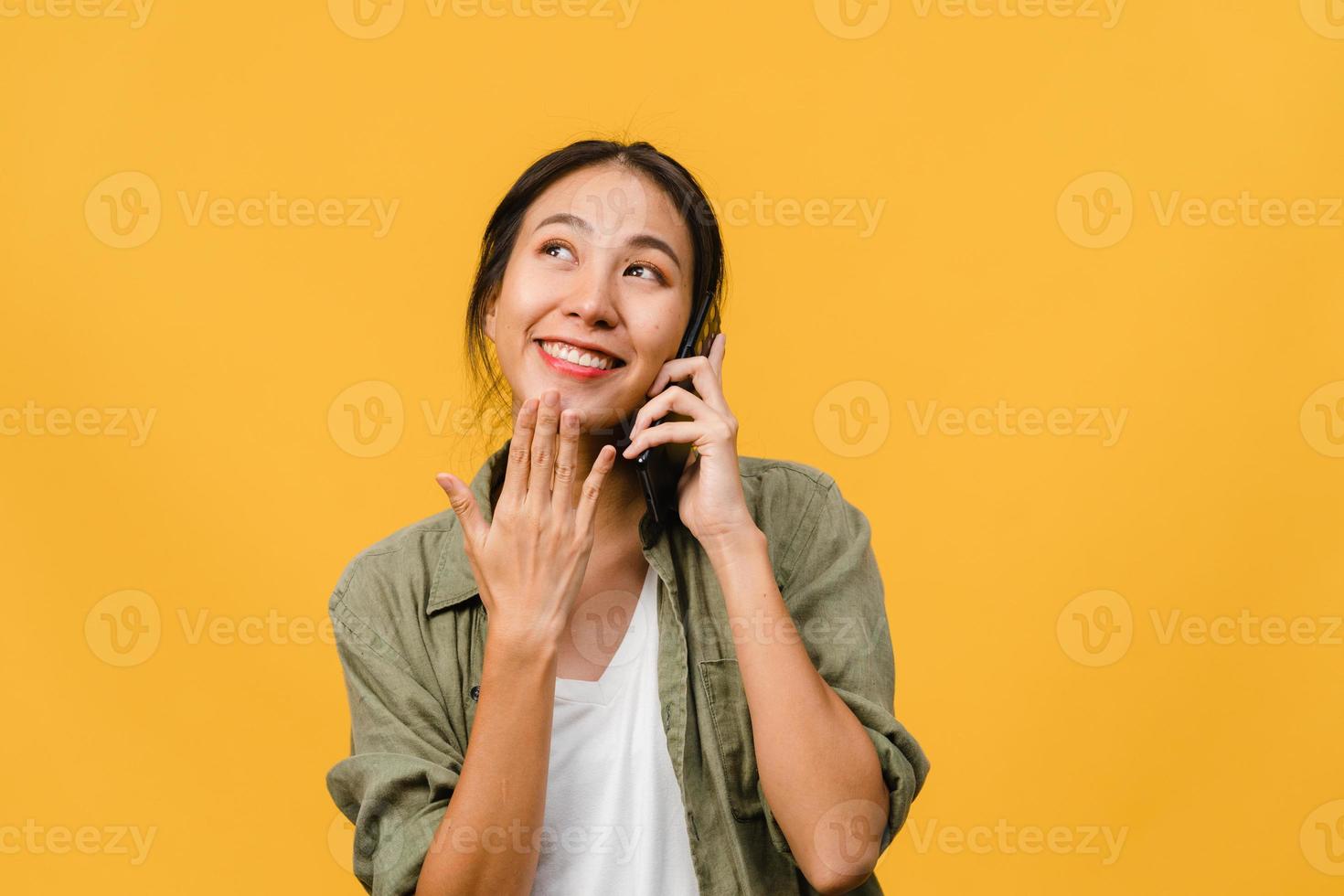 ung asiatisk dam pratar i telefon med positivt uttryck, ler brett, klädd i vardagskläder som känner lycka och står isolerad på gul bakgrund. glad förtjusande glad kvinna jublar över framgång. foto