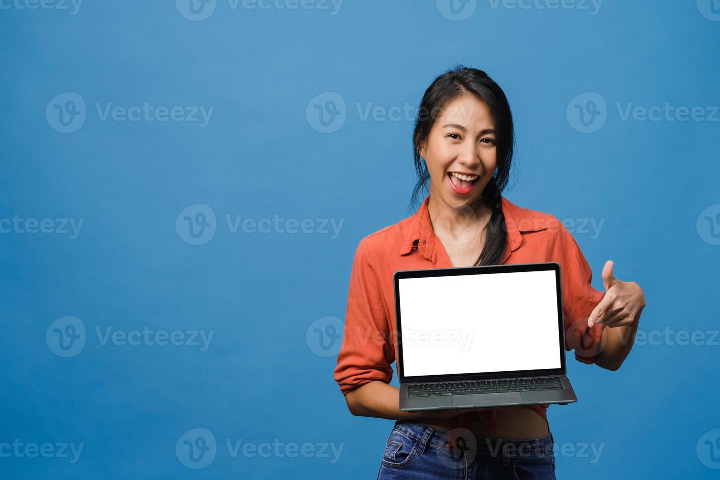 ung asiatisk dam visar tom bärbar datorskärm med positivt uttryck, ler brett, klädd i vardagskläder som känner lycka isolerad på blå bakgrund. dator med vit skärm i kvinnlig hand. foto