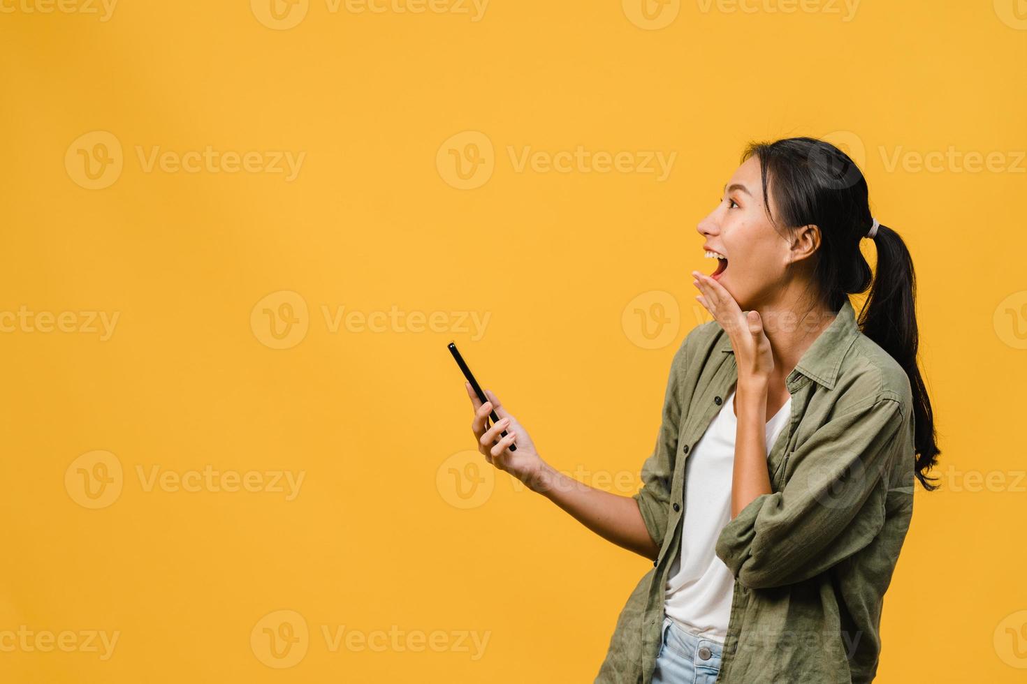 förvånad ung asiatisk dam med mobiltelefon med positivt uttryck, ler brett, klädd i vardagskläder och står isolerad på gul bakgrund. glad förtjusande glad kvinna jublar över framgång. foto