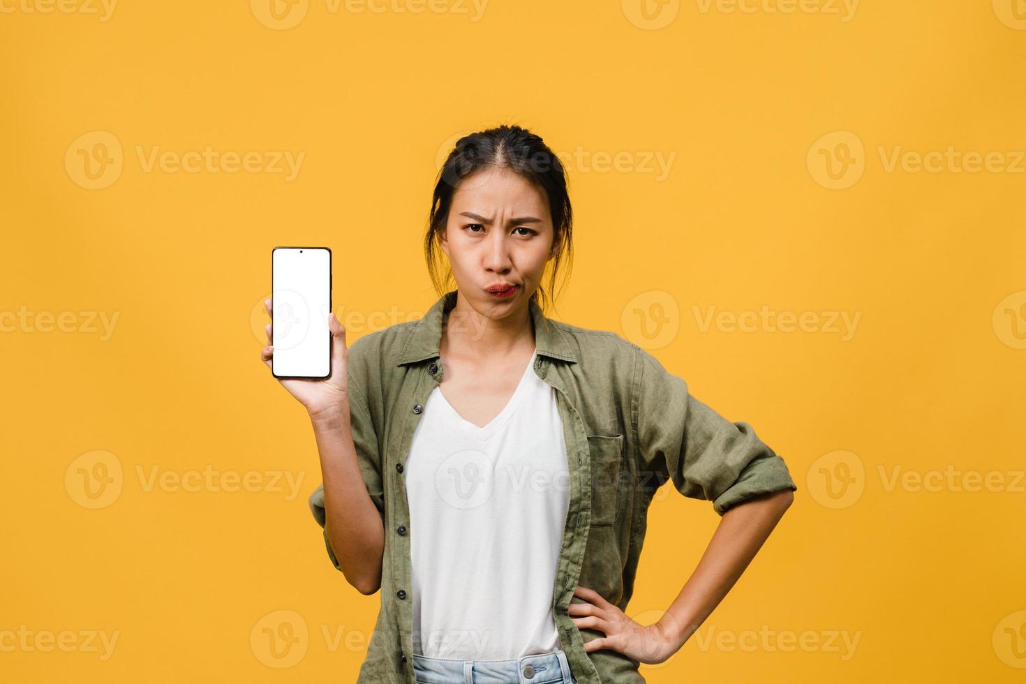 ung asiatisk dam visar tom smartphone -skärm med positivt uttryck, ler brett, klädd i vardagskläder som känner lycka på gul bakgrund. mobiltelefon med vit skärm i kvinnlig hand. foto