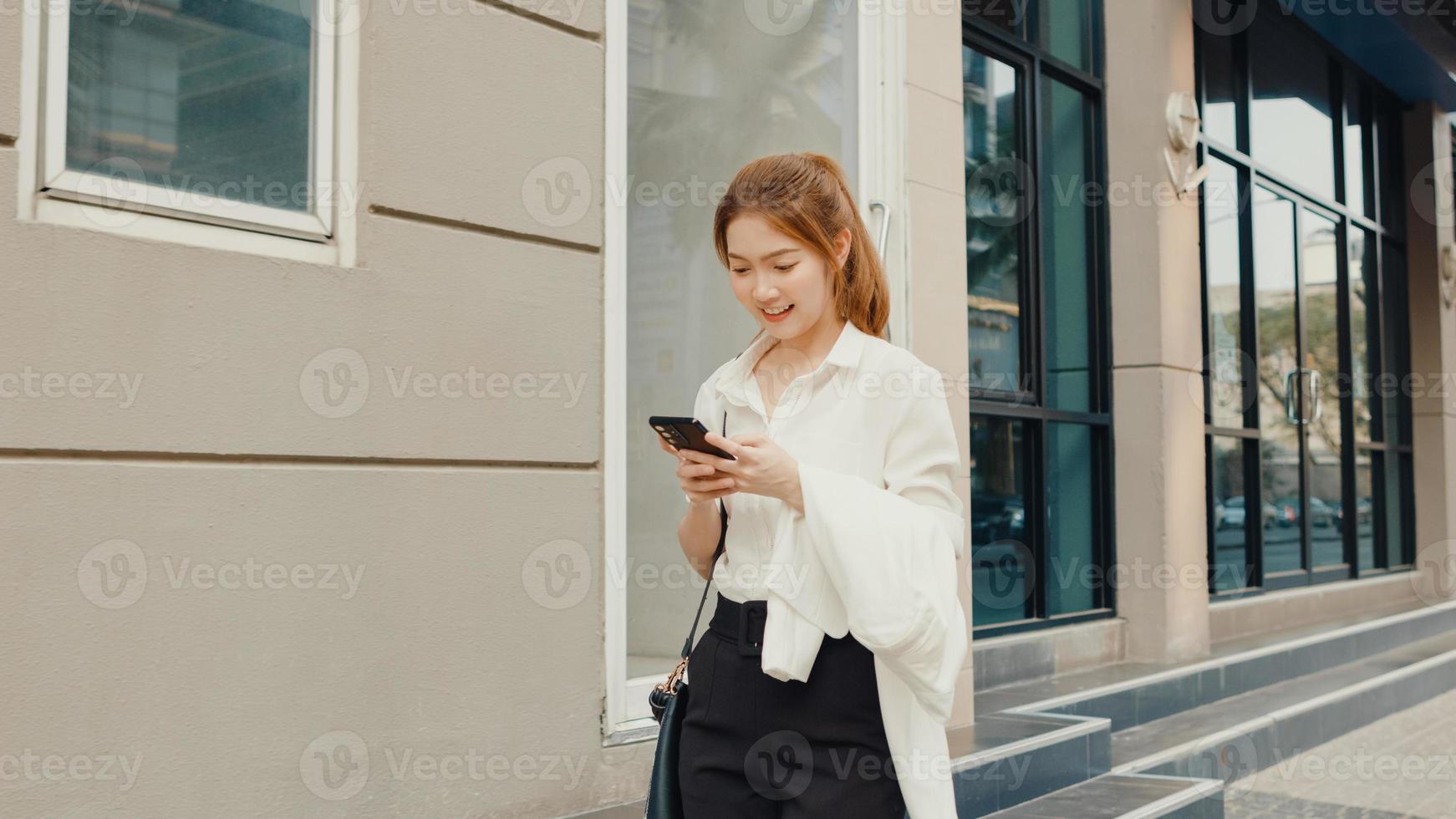 framgångsrik ung asiatisk affärskvinna i mode kontorkläder med smart telefon och skriver textmeddelande medan du går ensam utomhus i modern urban stad på morgonen. business on the go -koncept. foto