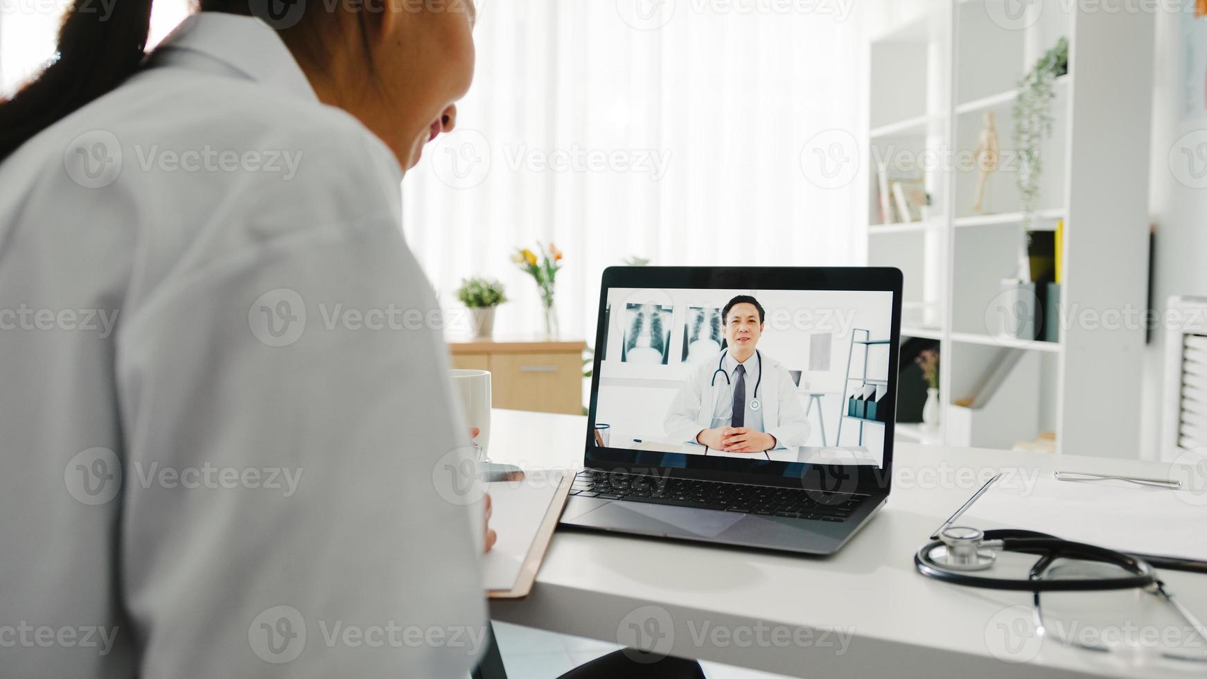 ung asiatisk damläkare i vit medicinsk uniform med bärbar dator som talar videokonferenssamtal med överläkare vid skrivbordet på vårdkliniken eller sjukhuset. social distansering, karantän för corona. foto