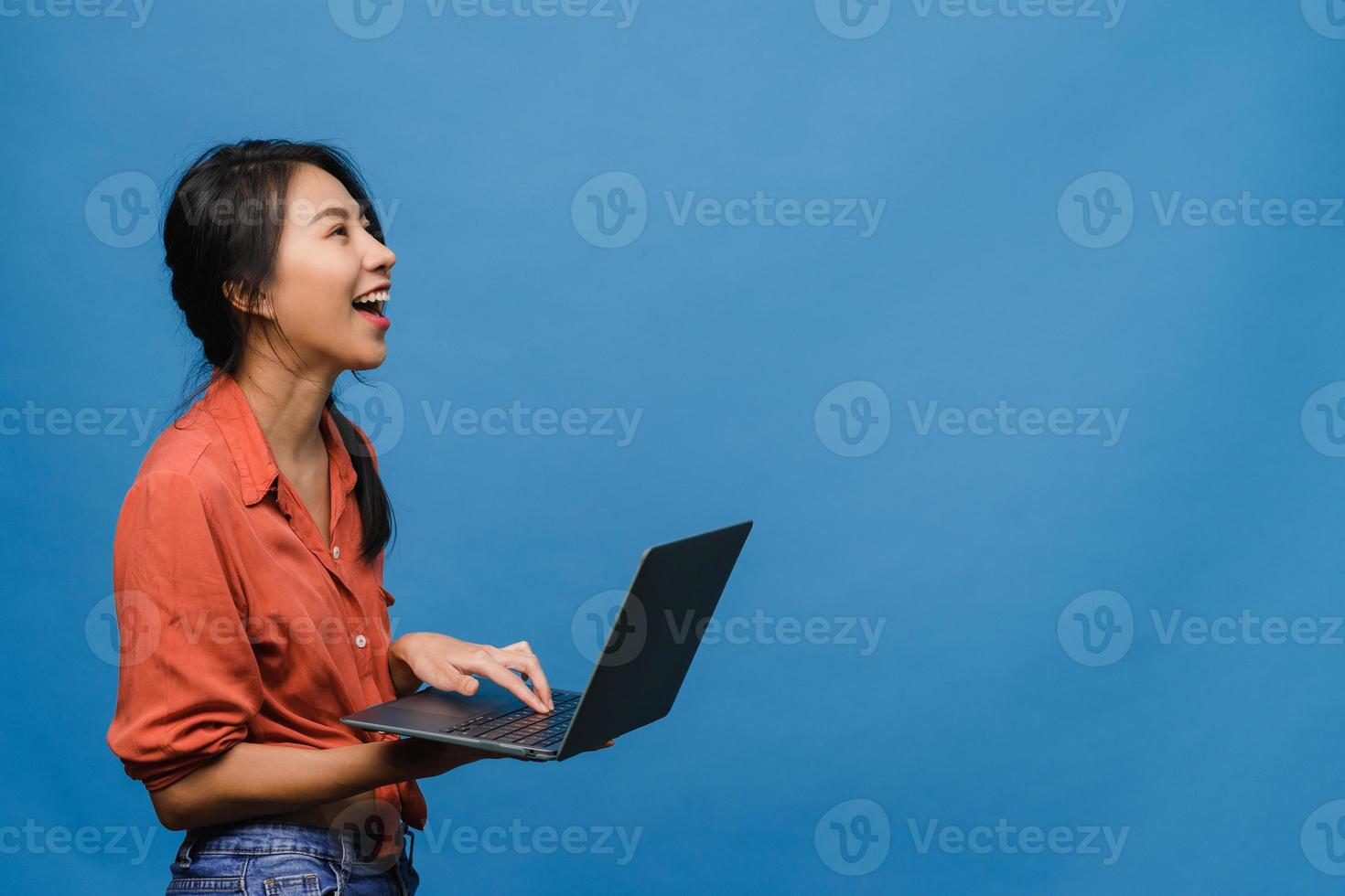 ung asiatisk dam som använder bärbar dator med positivt uttryck, ler brett, klädd i vardagskläder som känner lycka och står isolerad på blå bakgrund. glad förtjusande glad kvinna jublar över framgång. foto