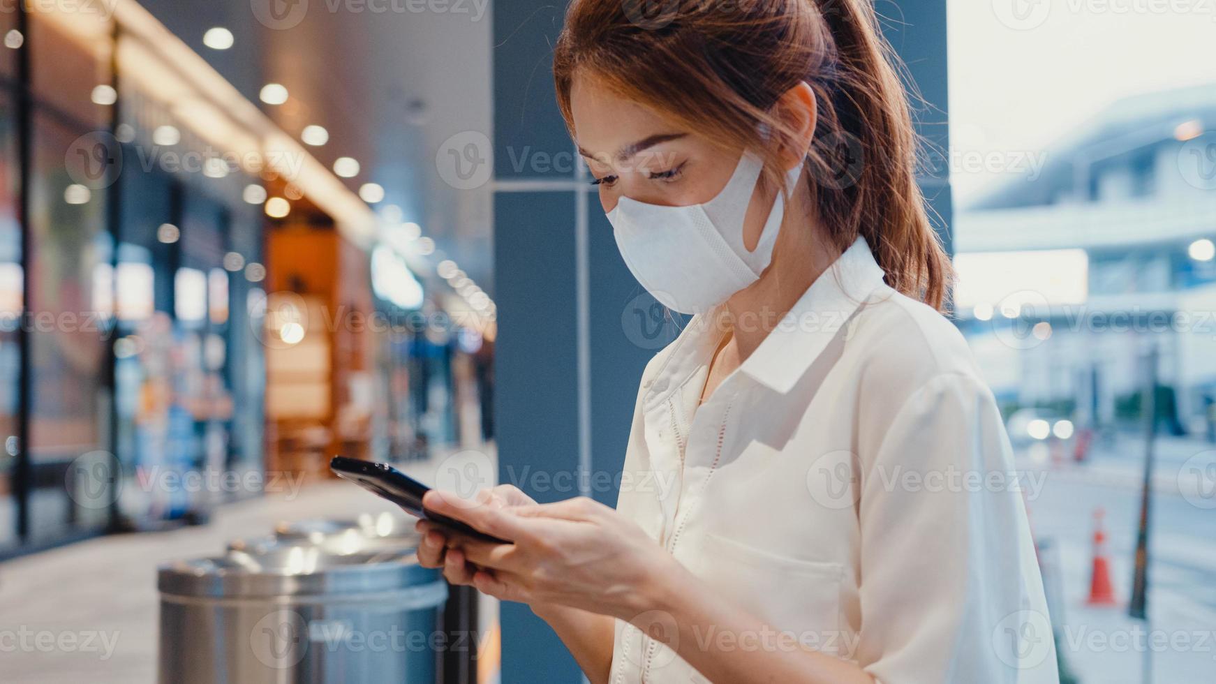 ung asiatisk affärskvinna i mode kontorkläder bär medicinsk ansiktsmask med smart telefon som skriver textmeddelande medan du sitter utomhus i en urban modern stad på natten. business on the go -koncept. foto