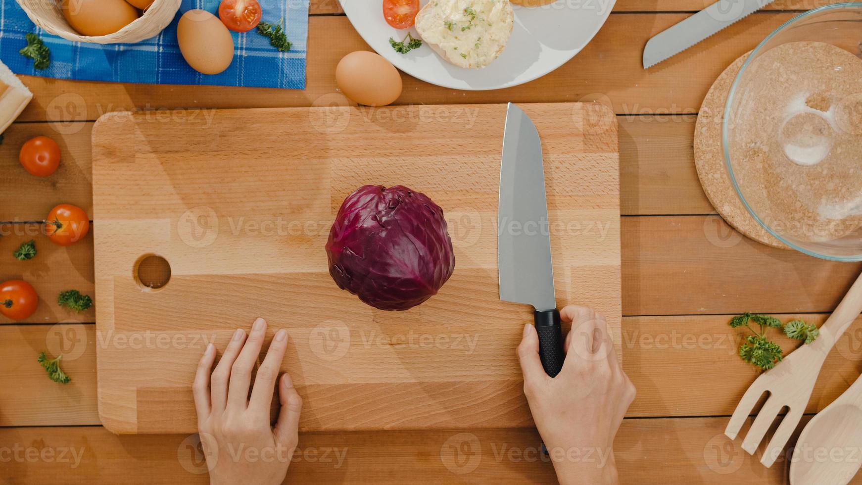 hand av ung asiatisk kvinna kock hålla kniv som skär rödkål på träskiva på köksbord i huset. att laga grönsaksallad, hälsosam mat och traditionella naturliga koncept. skott ovanifrån. foto