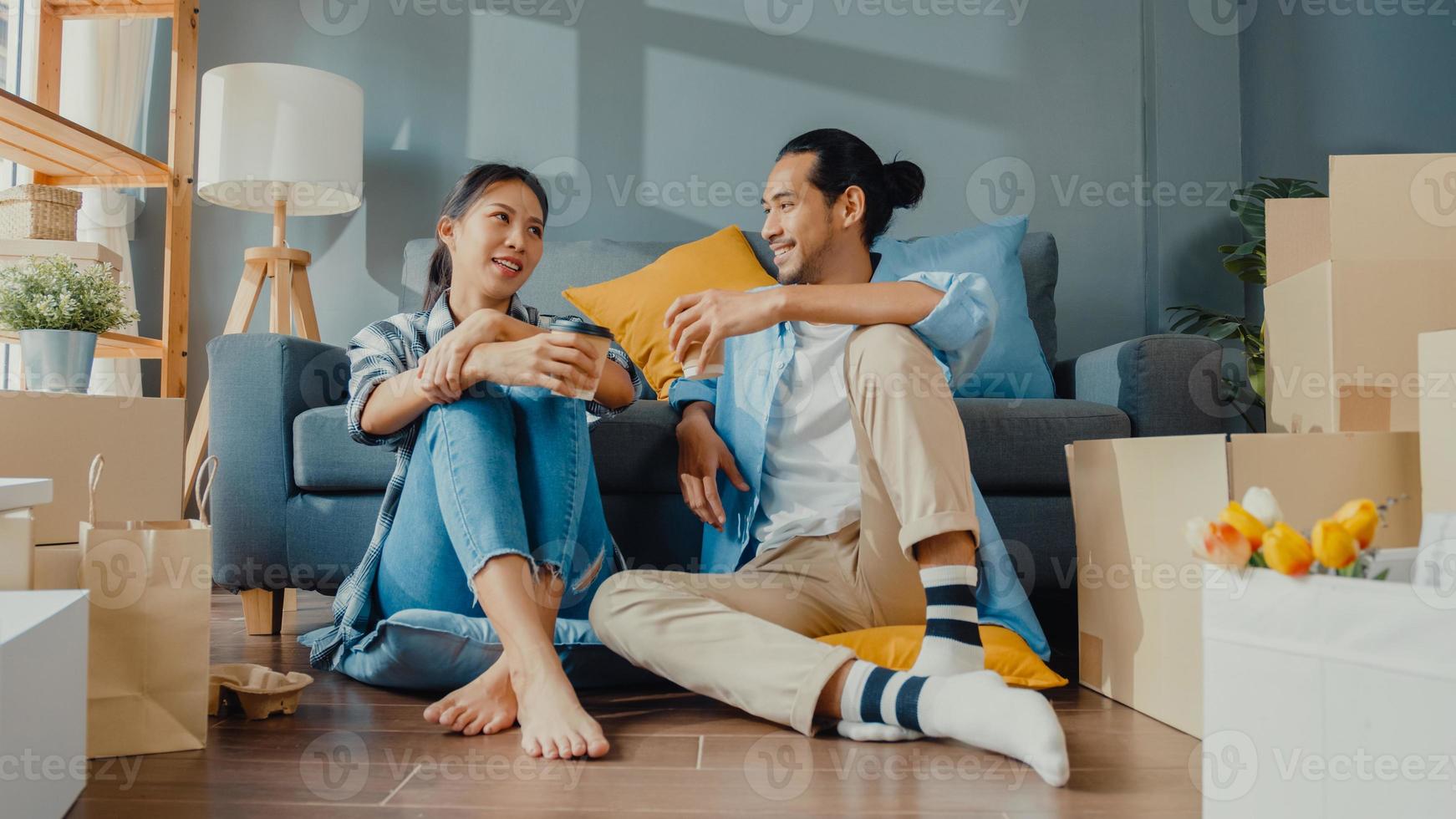 lyckliga asiatiska unga attraktiva par man och kvinna sitter vid nytt hem dricka kaffe koppla av och prata leende med kartongförpackning förvaring för att flytta i nytt hus. unga gifta asiatiska flytta hem koncept. foto