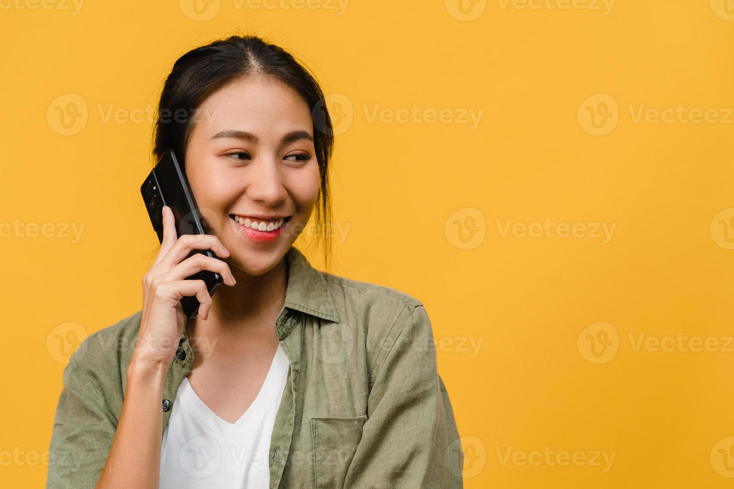 ung asiatisk dam pratar i telefon med positivt uttryck, ler brett, klädd i vardagskläder som känner lycka och står isolerad på gul bakgrund. glad förtjusande glad kvinna jublar över framgång. foto