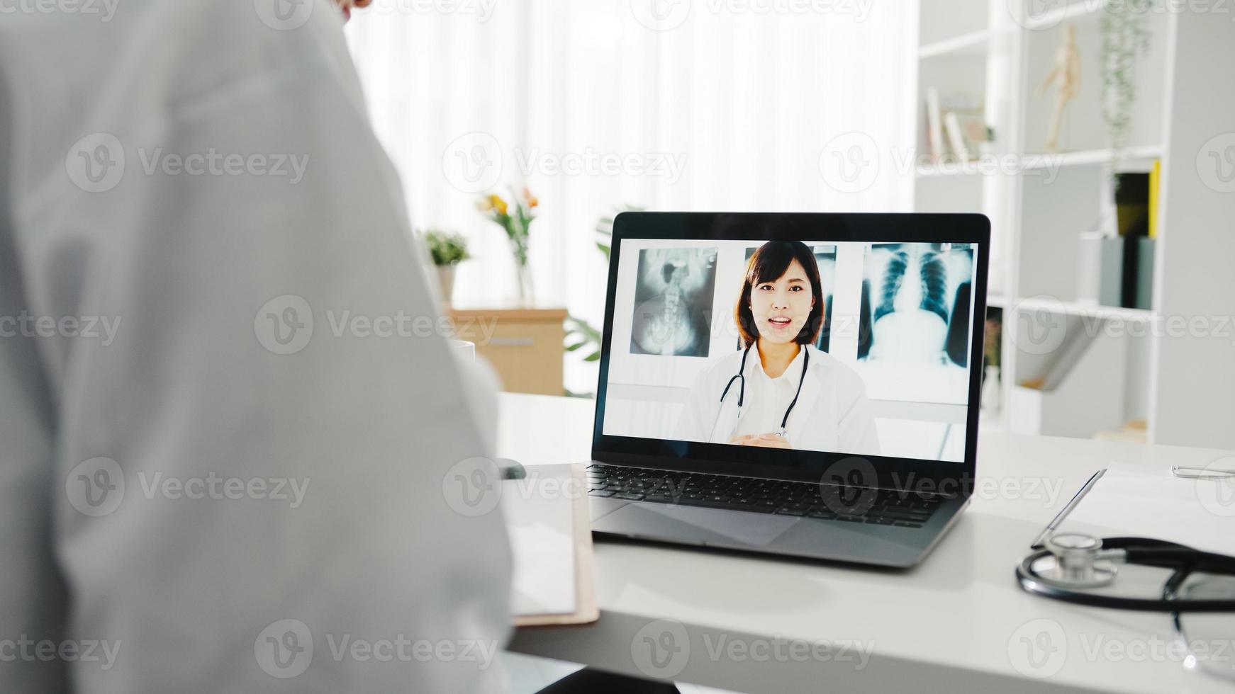 ung asiatisk damläkare i vit medicinsk uniform med bärbar dator som talar videokonferenssamtal med överläkare vid skrivbordet på vårdkliniken eller sjukhuset. social distansering, karantän för corona. foto