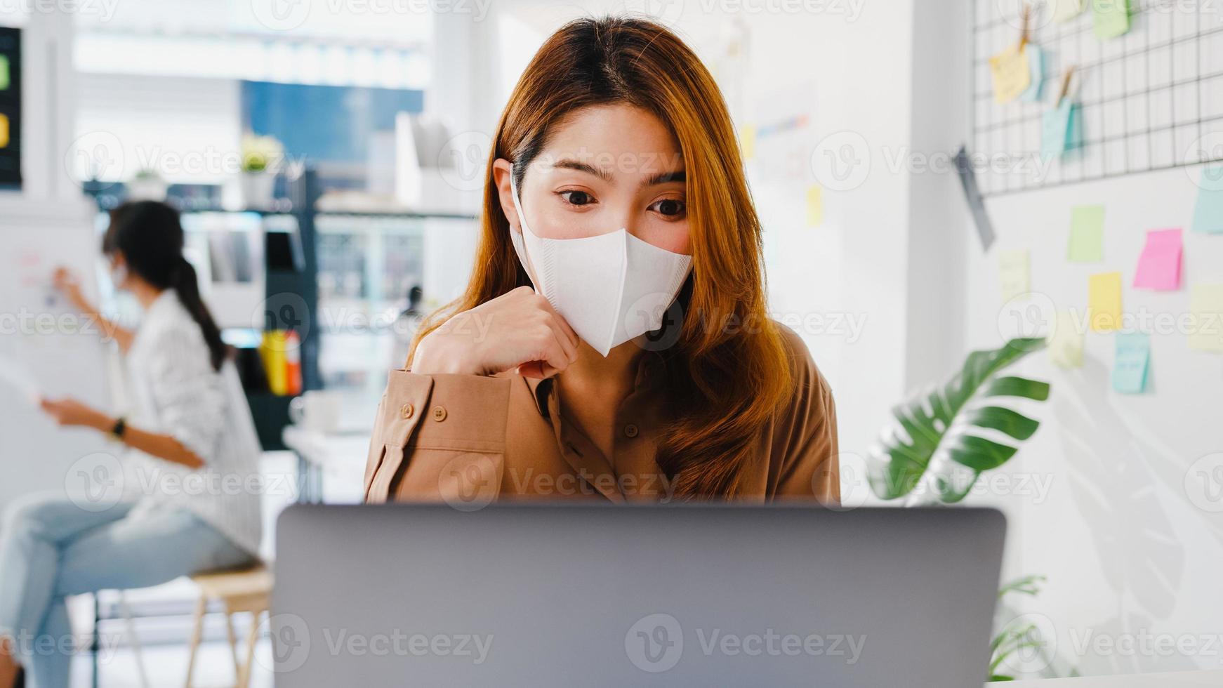 asiatisk affärskvinna bär ansiktsmask för social distansering i en ny normal situation för att förebygga virus medan du använder en bärbar datorpresentation för kollegor om planering i videosamtal medan du arbetar på kontoret. foto