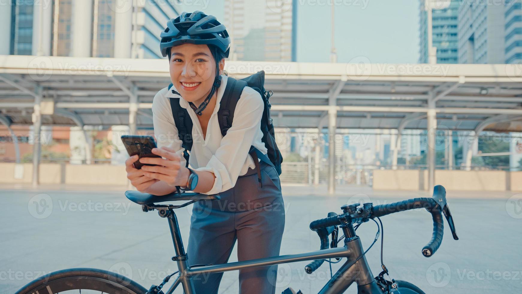 le asiatisk affärskvinna med ryggsäck använda smart telefon titta kamera i stad stå på gatan med cykel gå till jobbet på kontoret. sportflicka använder telefonen för arbete. pendla till jobbet, affärspendlare i staden. foto