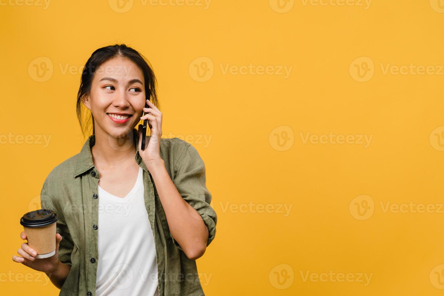 ung asiatisk dam pratar i telefon och håller kaffekoppen med positivt uttryck, ler brett, klädd i avslappnad trasa som känner lycka och står isolerad på gul bakgrund. ansiktsuttryck koncept. foto